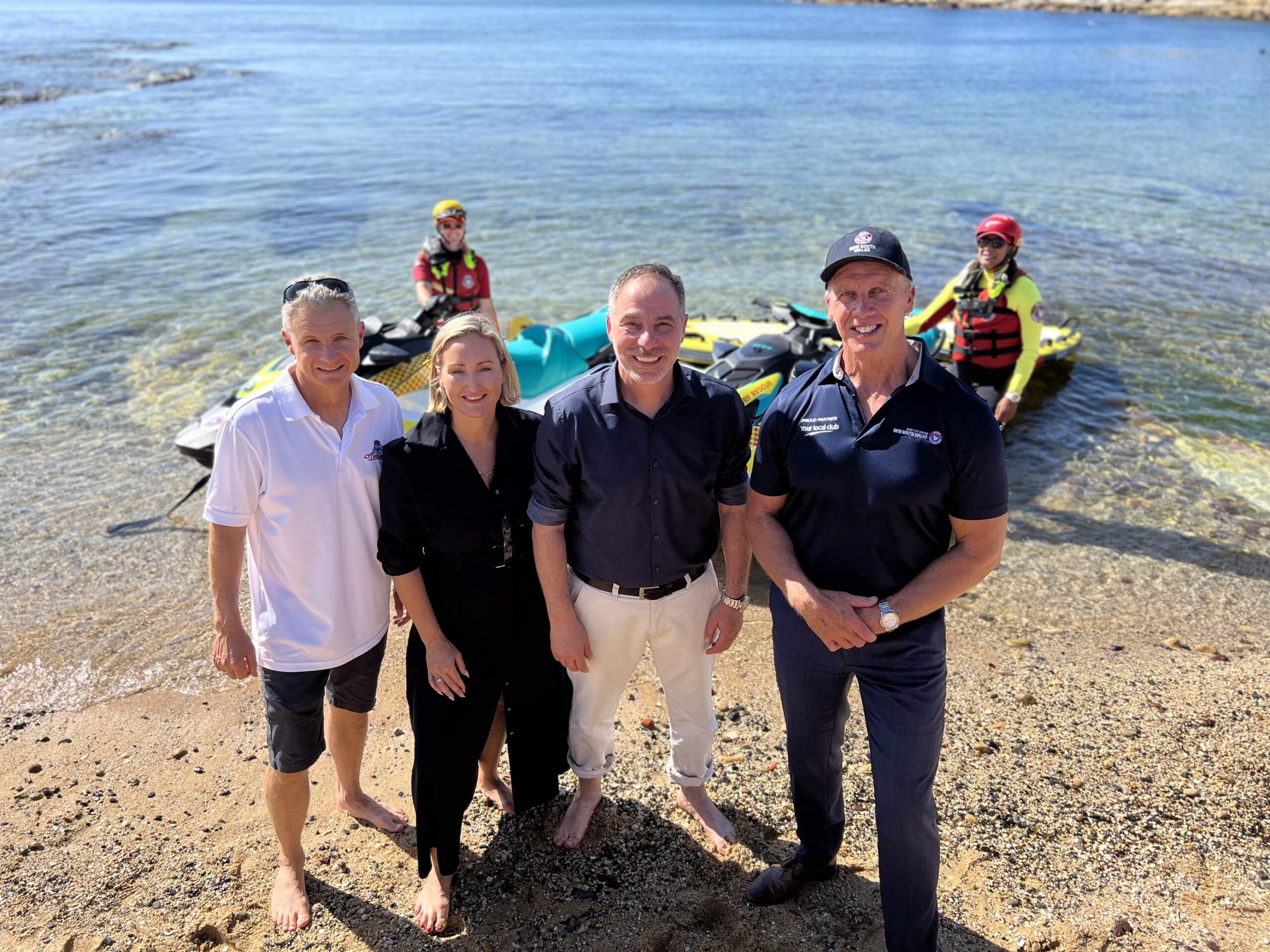 Minister for Emergency Services Jihad Dib2, Member for Coogee Dr Marjorie O’Neill, Federal Member for Kingsford Smith Matt Thistlethwaite and SLSNSW CEO Steve Pearce standing in front of two jetskis, operated by two female drivers.