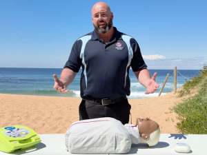 CPR trainer Dan Gay on a beach standing behind a CPR manikin