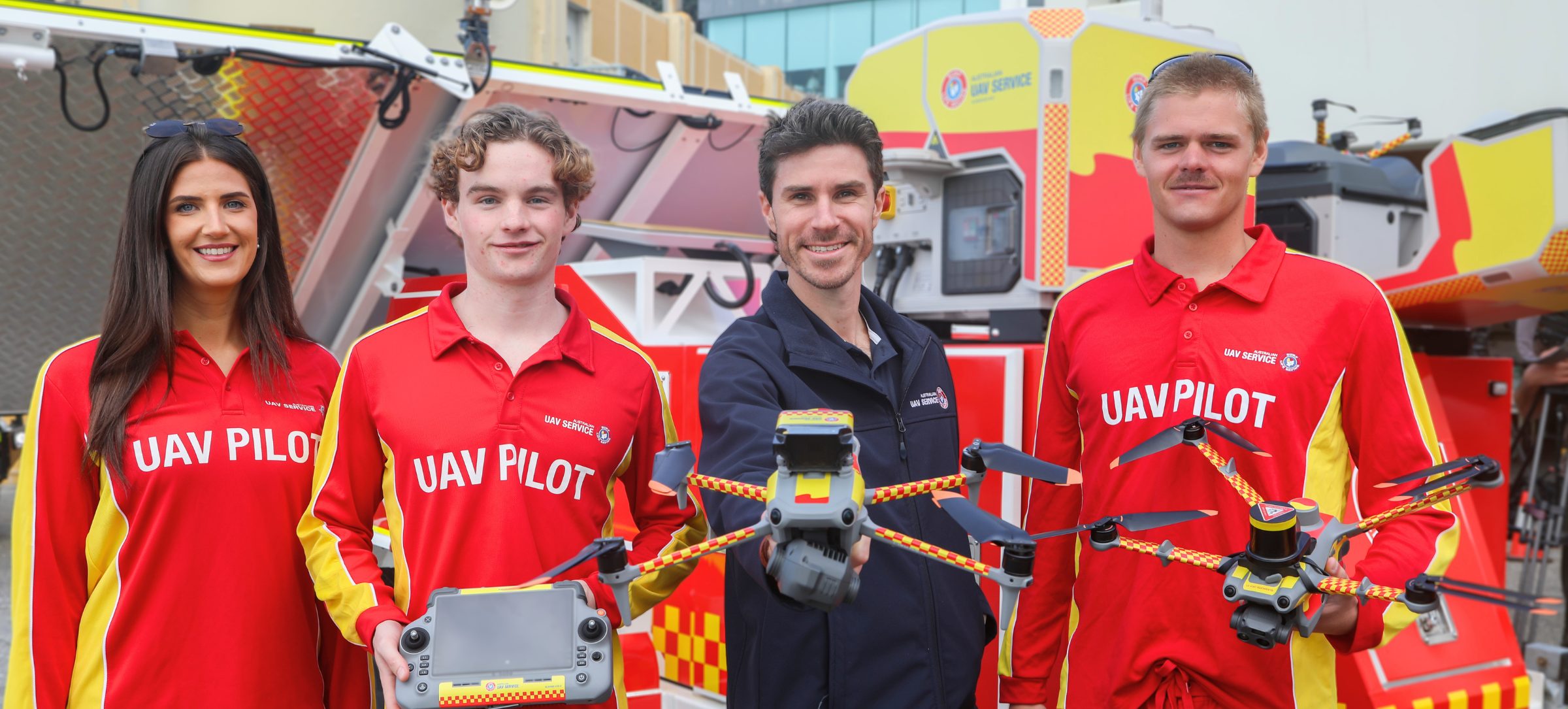 Four UAV service team members in red “UAV PILOT” uniforms stand in front of an emergency response vehicle, holding drones and a remote controller.